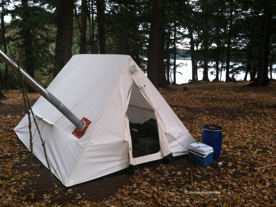 Snowtrekker 10x13 Crew setup in clearing next to lake shore with trees between door is open into the tent stove pipe is exiting left side of tent supported by two sticks