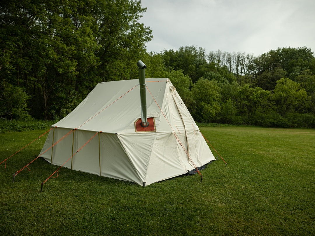 Snowtrekker 10x13 Outfitter setup in green grass tress in background left side View Stove Pipe Exiting Tent With Stove Pipe Support Ring holding the stove pipe with three orange guy lines for support in a tripod configuration