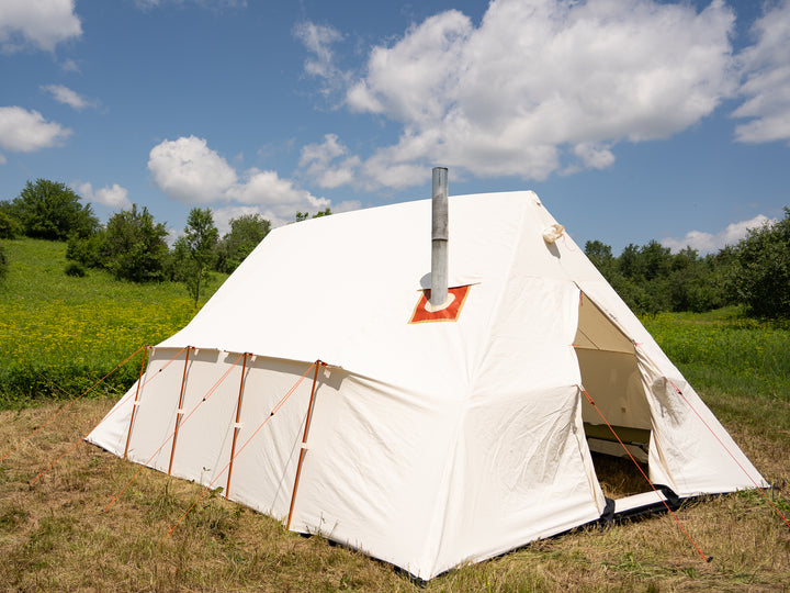 White canvas tent with a chimney on a grassy field under a blue sky with clouds.