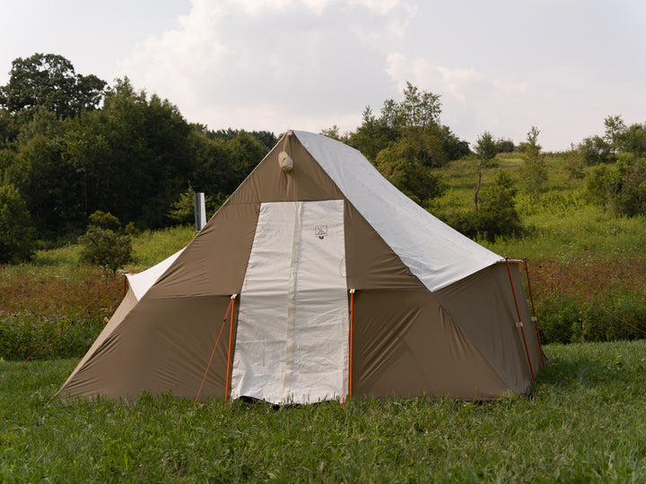 Brown nylon and white canvas camping tent with a white door in a grassy field with trees in the background