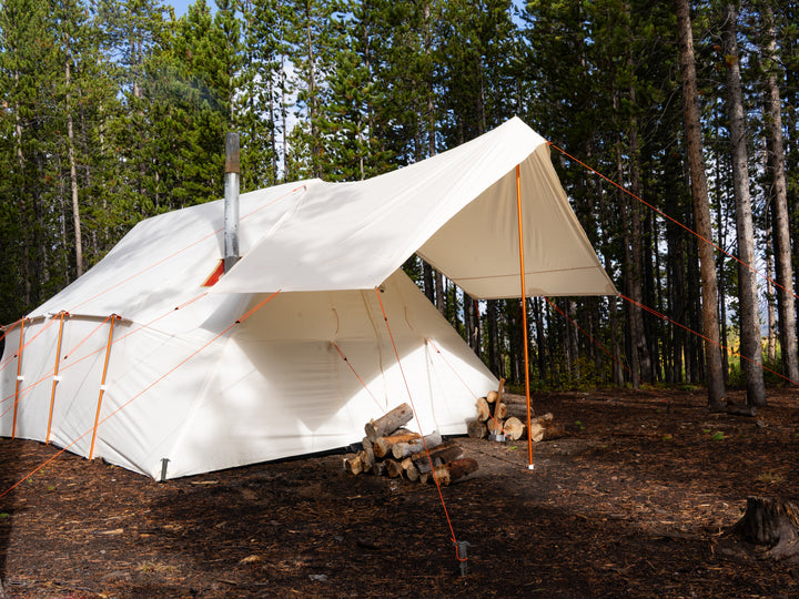 White canvas camping tent with awning and wooden fire logs in a forest setting