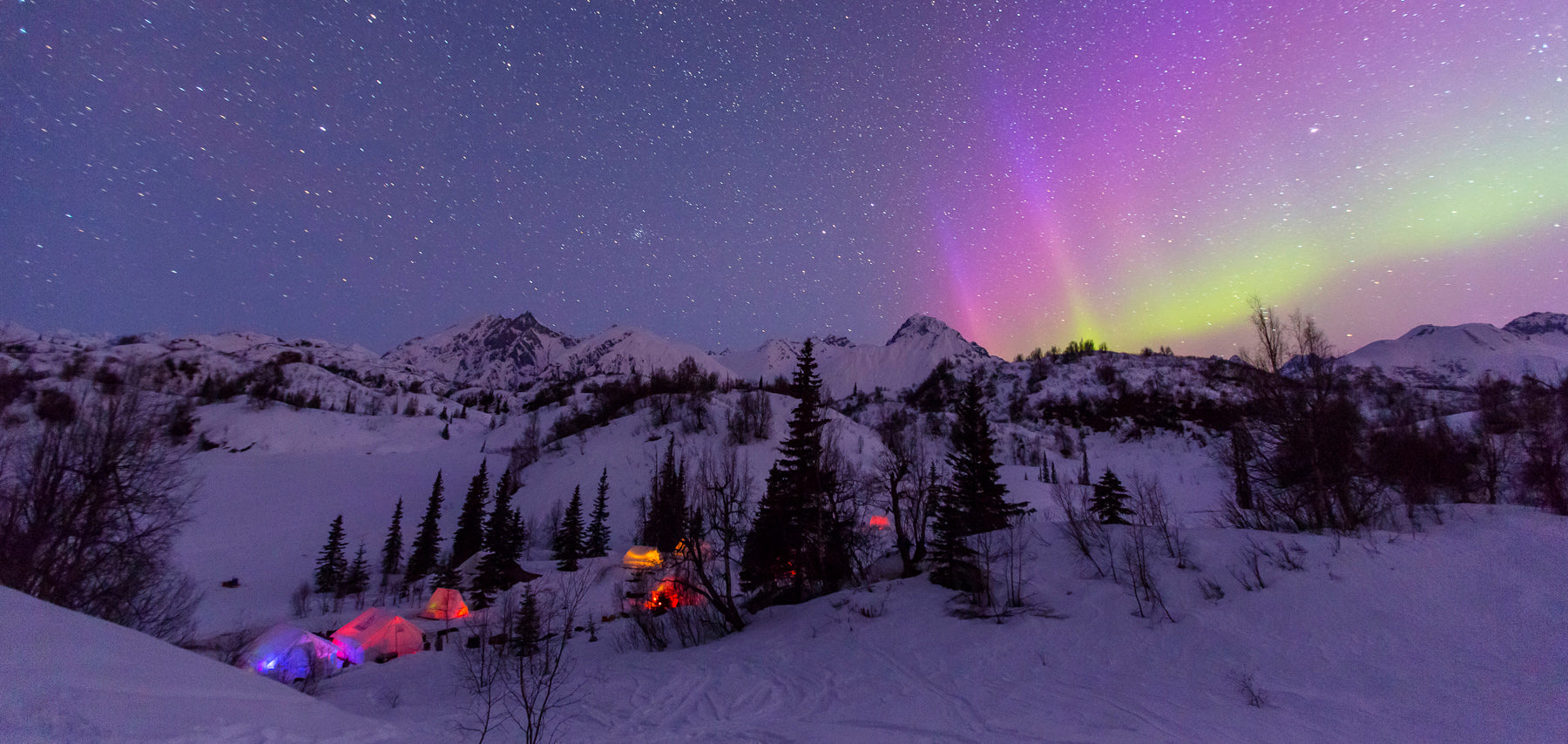 Winter mountain scene at night with the northern lights and several lit tents