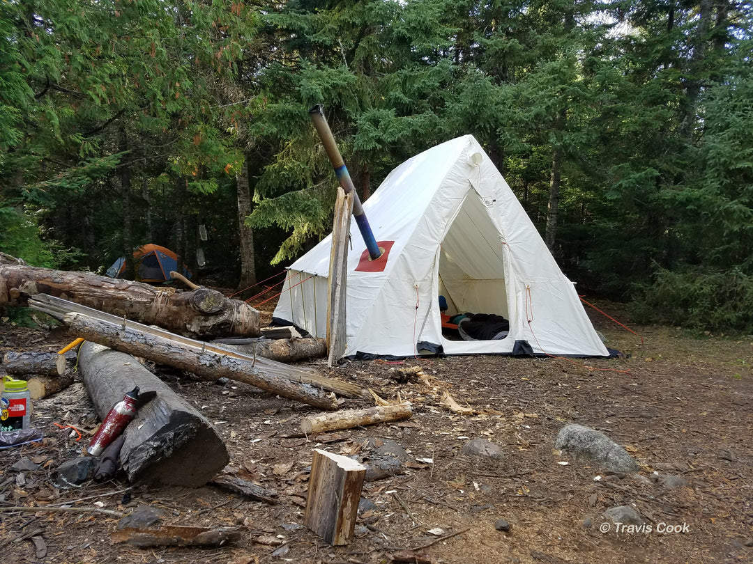 Snowtrekker 9x11.5 Basecamp Setup Door Open sleeping pad in tent along right sidewall stove pipe exiting left side of tent propped up with  log next logs on the ground with tent and pine trees in background