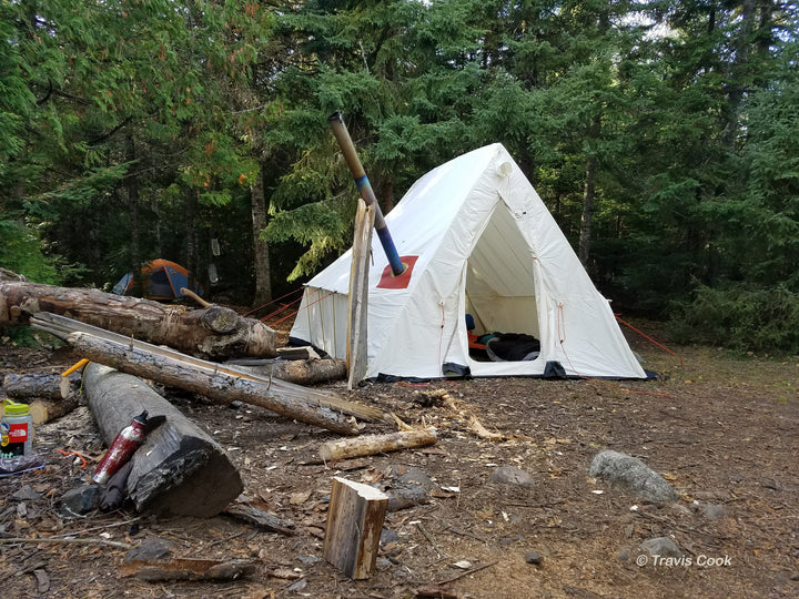 Snowtrekker 9x11.5 Basecamp Setup Door Open sleeping pad in tent along right sidewall stove pipe exiting left side of tent propped up with  log next logs on the ground with tent and pine trees in background