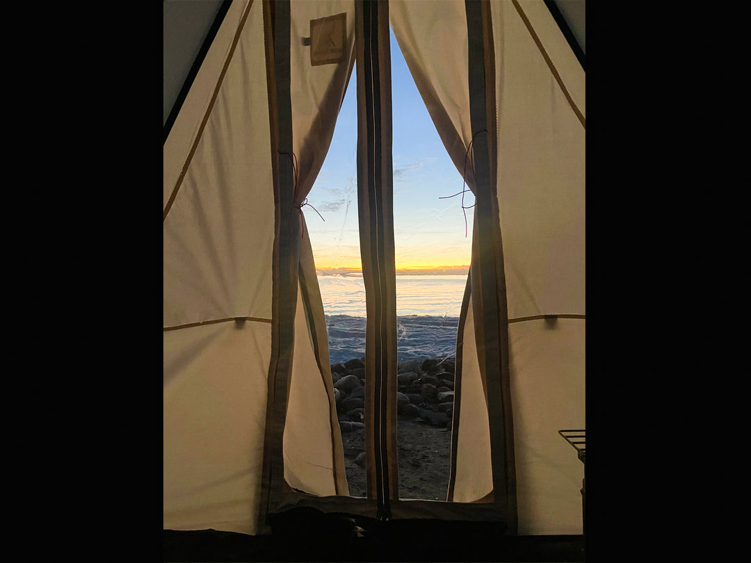 Looking through clear door from inside of a Snowtrekker tent at a frozen lake with sun setting