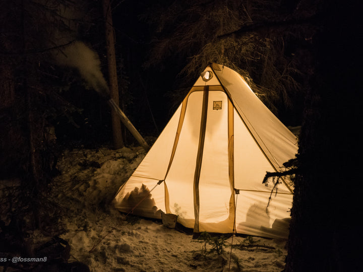 Snowtrekker 8x10 Crew Setup In Winter at Night With Lights In Tent stove pipe exiting left side of tent with smoke coming out snow on the ground and trees in background