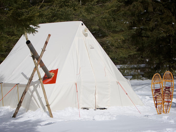 Snowtrekker 9x11.5 Basecamp Setup In Winter snow on the ground stove chimney exiting the left side of the tent supported by sticks snowshoes standing up in snow on right side of tent trees in background