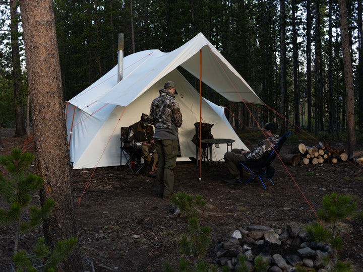 Three people camping in a forest with a large white canvas hot tent.