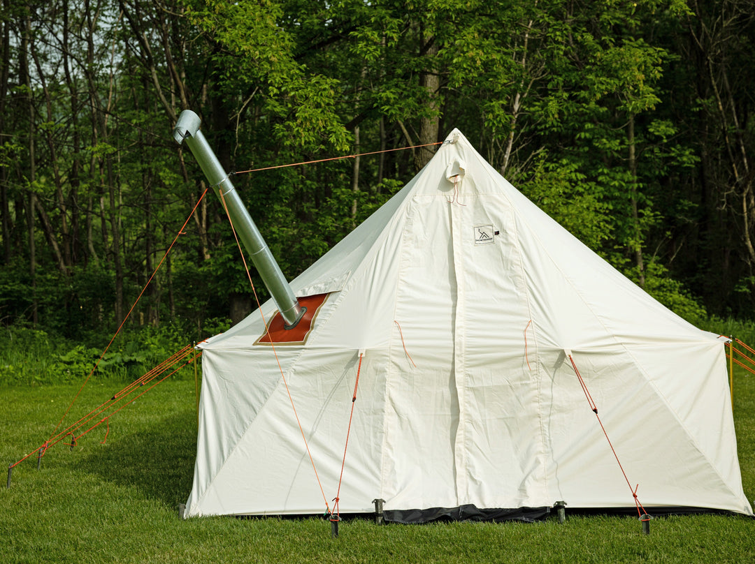 Snowtrekker 10x13 Outfitter looking at the front with door closed setup on green grass with tress in the background with Stove Pipe Support Ring On Chimney exiting the left side of the tent held in place by orange guy lines secured to the tent and tent stakes like a tripod