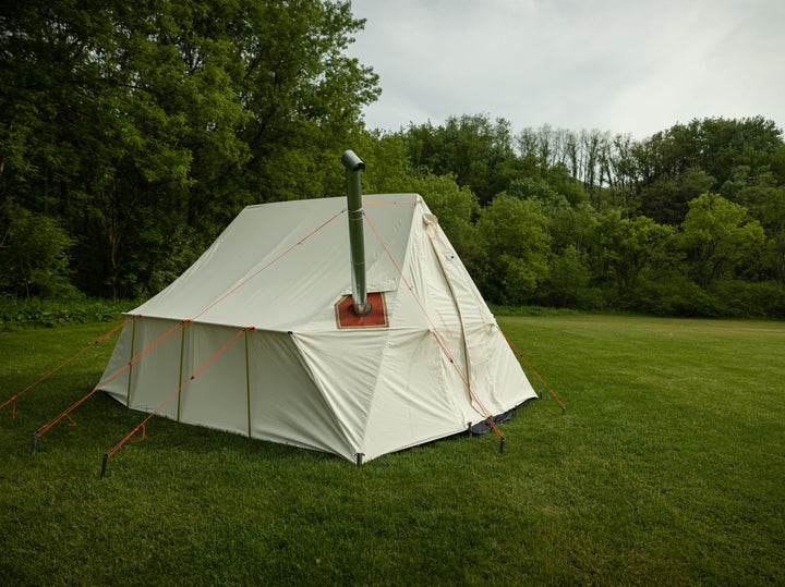 Snowtrekker 10x13 Outfitter setup in green grass tress in background left side View Stove Pipe Exiting Tent With Stove Pipe Support Ring holding the stove pipe with three orange guy lines for support in a tripod configuration