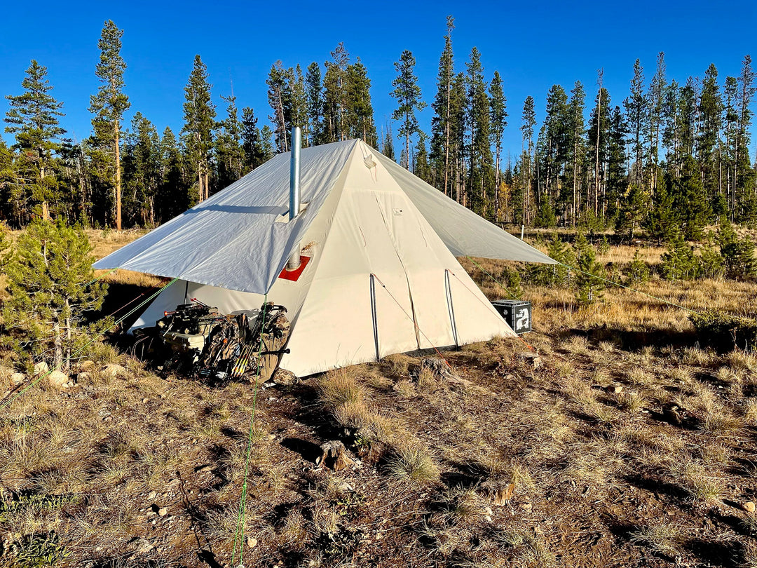 Snowtrekker High Country Tent WIth Fly archery hunting equipment stored under the fly on left side of tent pine trees in background in Colorado mountain meadow