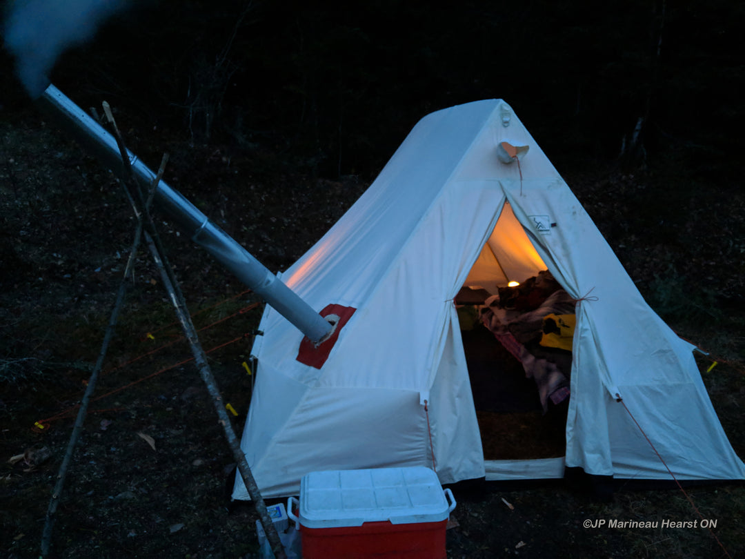 Snowtrekker 9x11.5 Shortwall With Door Open, Light In Tent cot along right sidewall and Smoke Coming Out of Stove Pipe on left side of tent propped up by sticks red cooler in front of tent