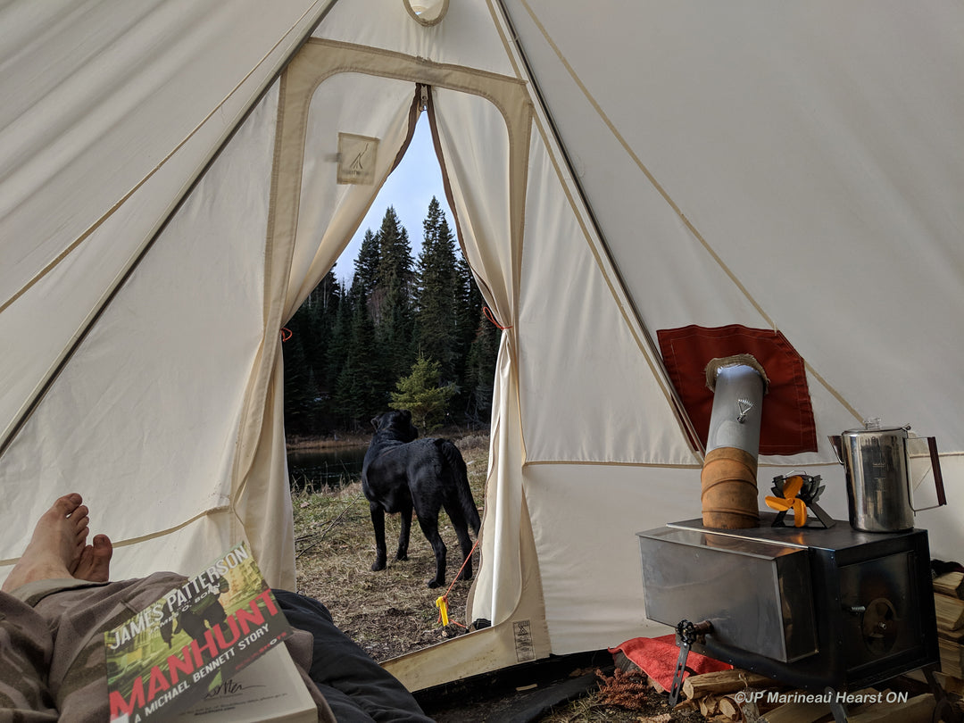 Snowtrekker 9x11.5 Shortwall Inside Tent Looking Out Open Door with person laying on cot feet visible stove with wood next to it on right side of tent coffee pot on stove