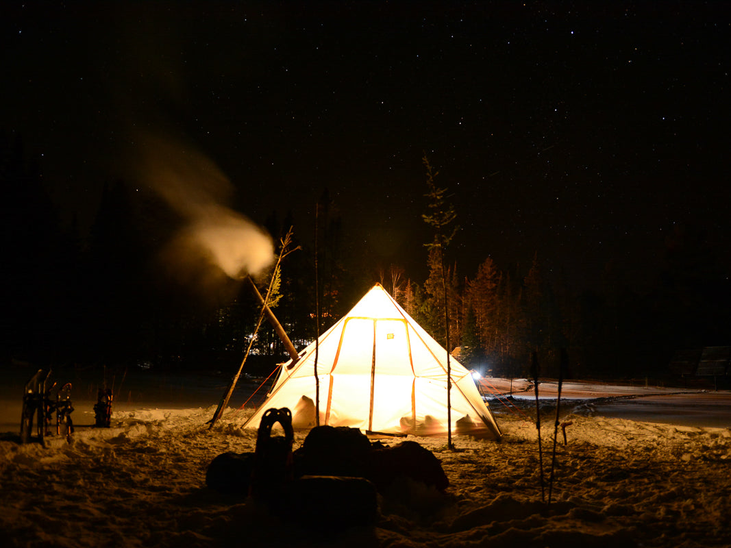 Snowtrekker 13x18 Mega Crew Setup In Winter at night with Lights In Tent Smoke Exiting Stove Pipe supported by sticks snowshoes in snowbank trees in background 