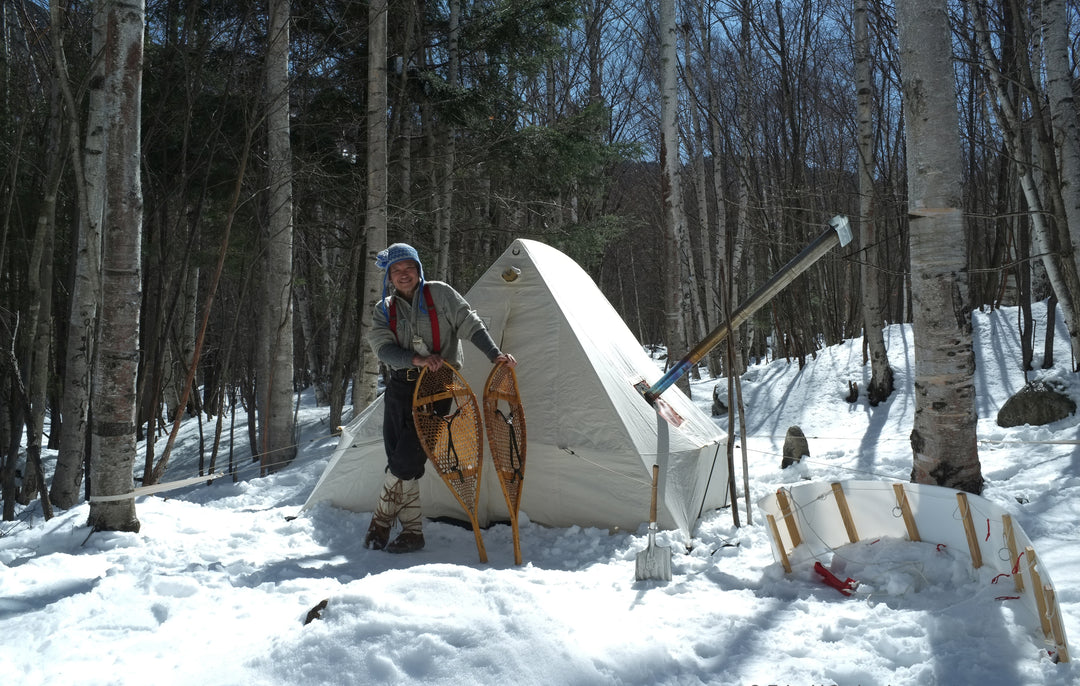 Snowtrekker 8x10 Shortwall Setup In Winter WIith Man Holding Snowshoes In Front Of Tent snow on the ground trees in background and toboggan to the right of the tent