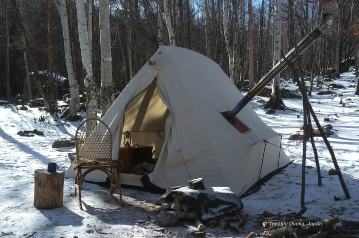 Snowtrekker 8x10 Shortwall Setup In Winter chair in front of tent next to wood for stove chimney exiting right of tent supported by sticks snow on the ground trees in background