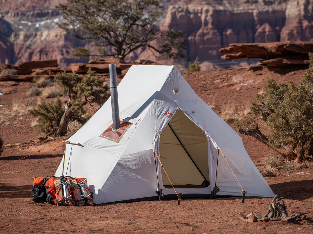 White camping tent with a chimney in a desert landscape with cliffs and sparse vegetation.