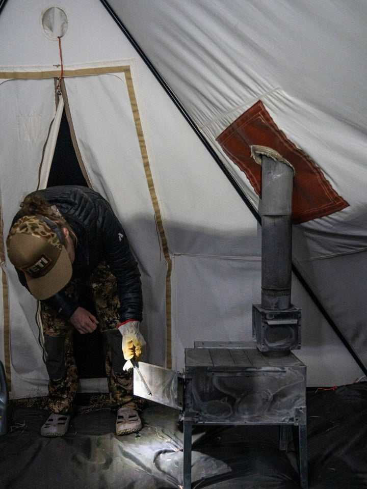 Person using a camping stove inside a tent with a visible chimney.
