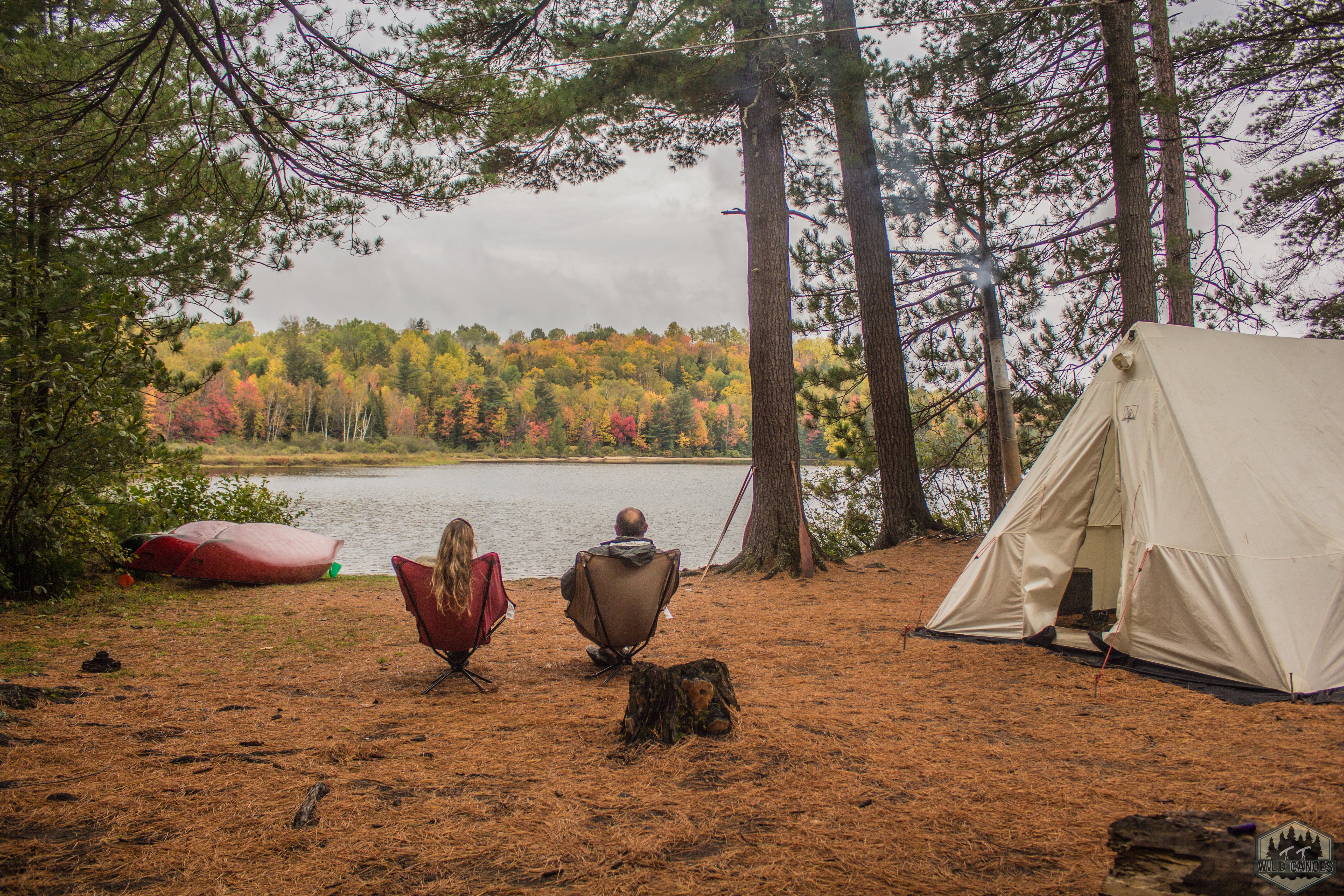 two people sitting in camp chairs near a tent at the edge of a lake